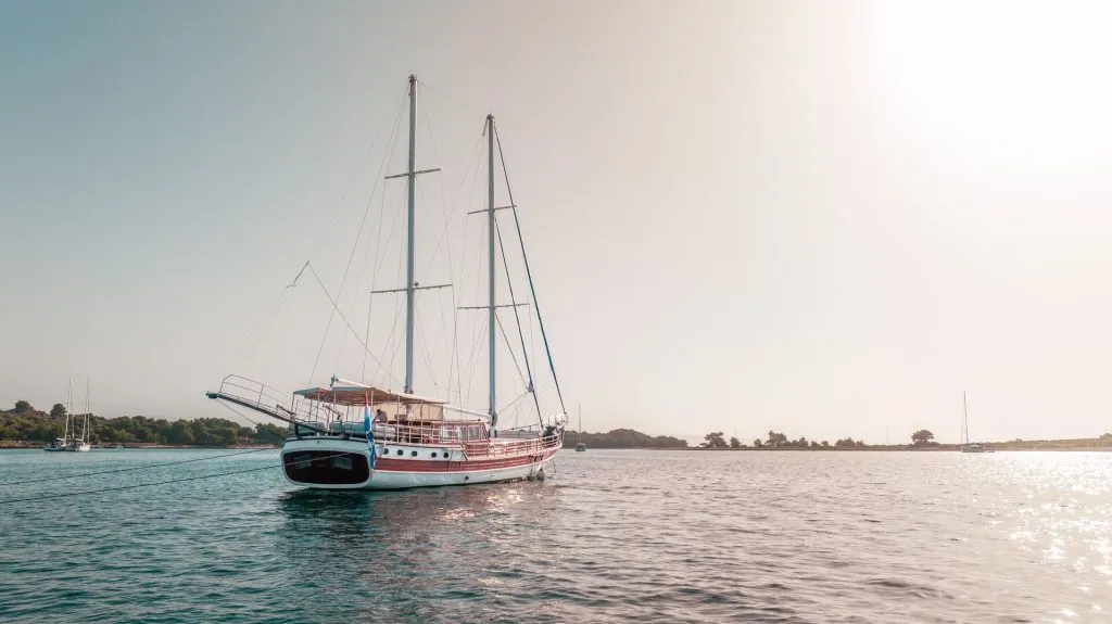 A sailboat with two masts floats on calm water near the shore under a clear sky, with sunlight illuminating the scene and a few trees visible in the background.