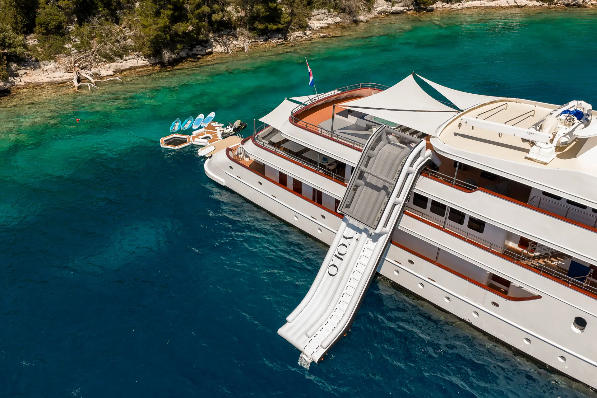 A luxury yacht anchored near a forested shoreline has a large inflatable slide extending into turquoise water, with kayaks and paddleboards floating nearby.