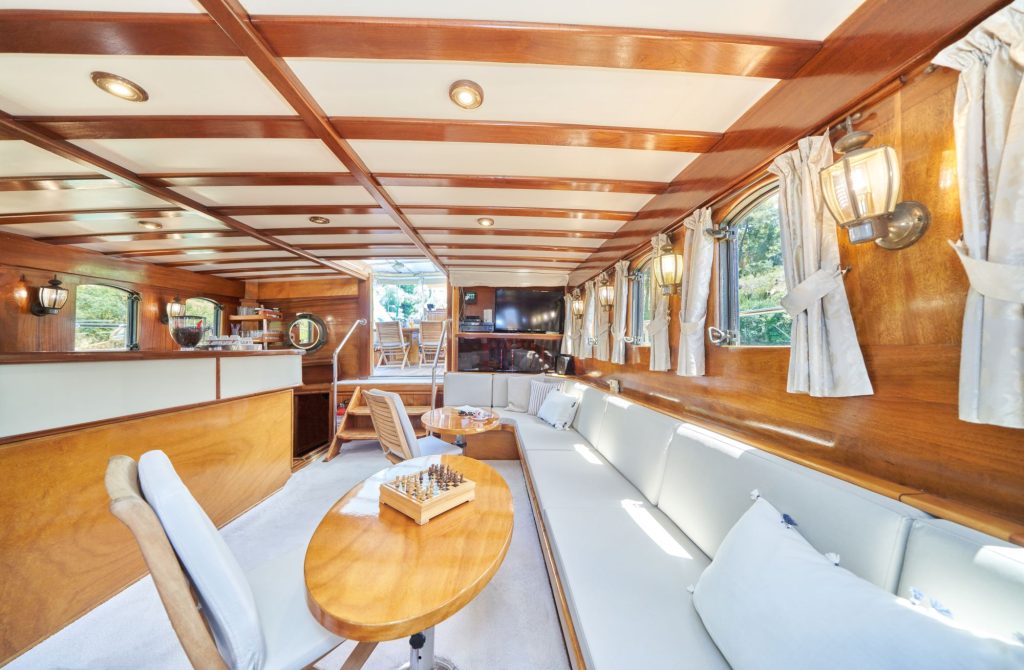 LIBRA Luxurious yacht interior with wood paneling, long cushioned seating, a round table with a chessboard, large windows with curtains, wall lamps, and an open dining area in the background. Natural light fills the space.