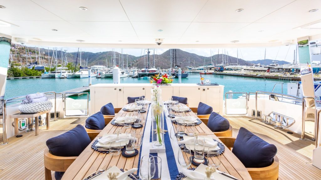 A luxurious yacht dining area set for a meal, featuring a long wooden table with place settings, navy cushions, flowers, and views of a marina, docked boats, and mountains in the background.