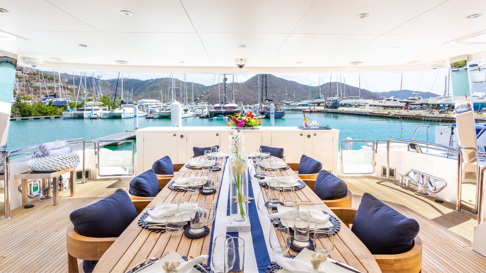 A luxurious yacht dining area set for a meal, featuring a long wooden table with place settings, navy cushions, flowers, and views of a marina, docked boats, and mountains in the background.