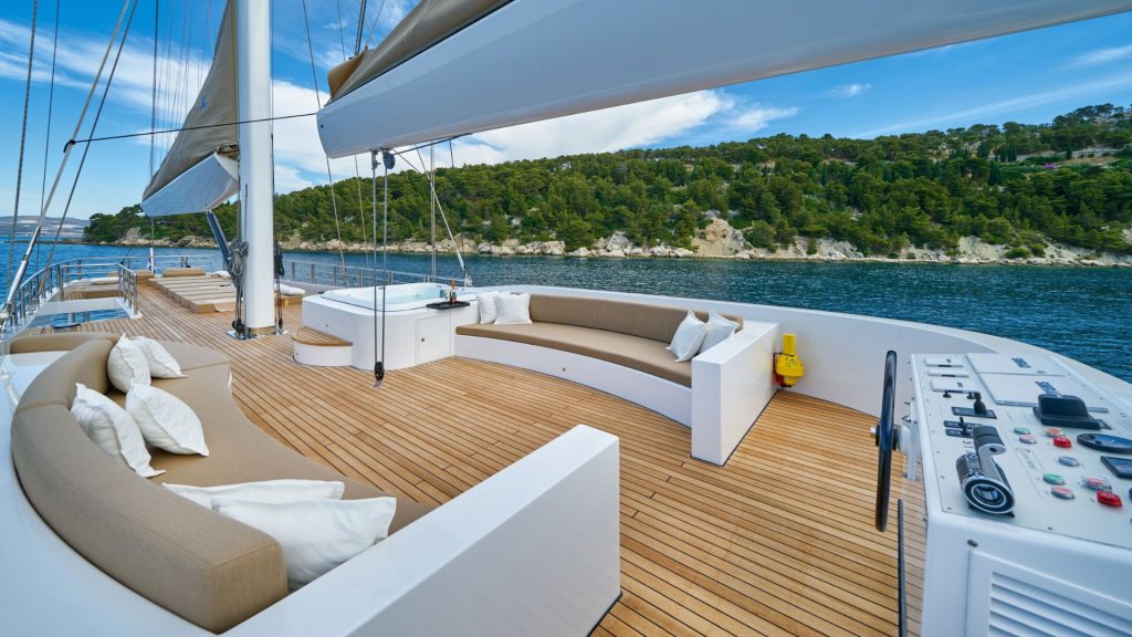 Spacious yacht deck with cushioned seating, a hot tub, and control panels; surrounded by blue water and forested shoreline under a partly cloudy sky.