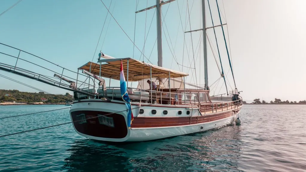 A wooden sailboat with a Dutch flag anchored on calm blue water, with a canopy covering the deck and green shoreline visible in the background under a clear sky.