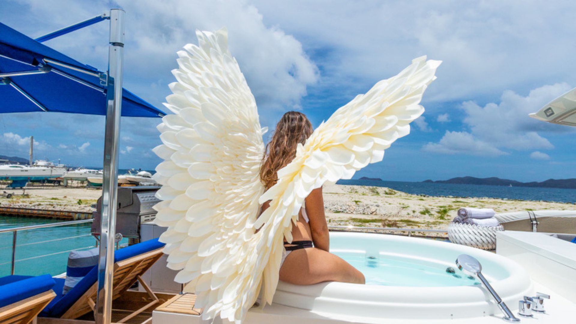 A woman wearing large, white angel wings sits in a hot tub on a yacht, facing the ocean and rocky shore under a partly cloudy sky. Lounge chairs and other yachts are visible nearby.