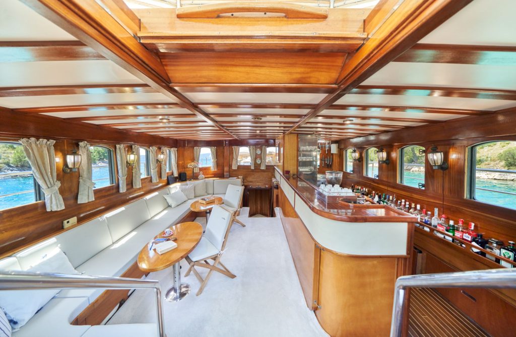 LIBRA Interior view of a luxurious yacht with polished wood paneling, cream-colored seating, a bar stocked with bottles, large windows with curtains, and a scenic view of the sea outside.