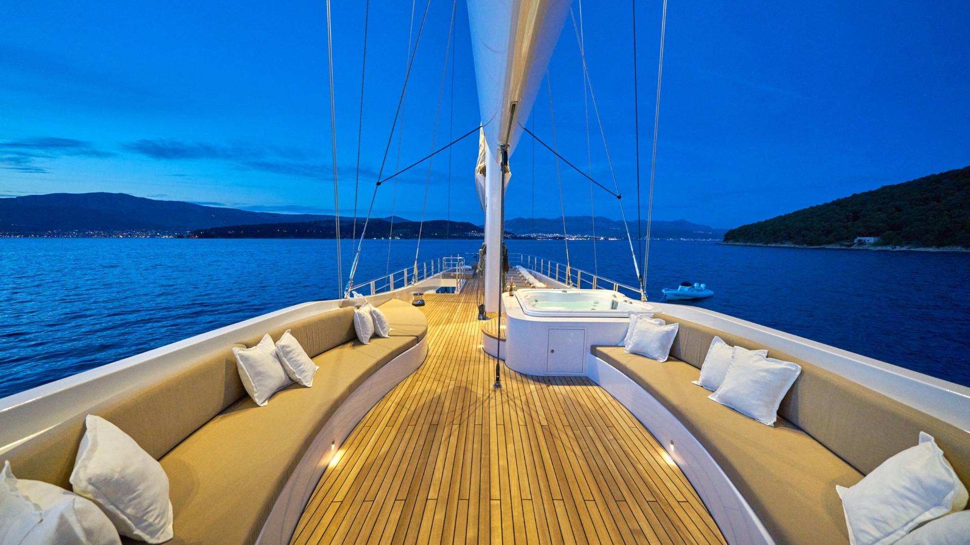 View from the deck of a luxury yacht at dusk, featuring curved seating with white pillows, a hot tub in the center, and calm blue water with distant shorelines and hills in the background.