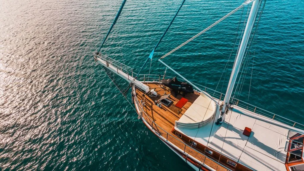 A top-down view of a sailboat with wooden decking and white sails floating on calm, deep blue water under bright sunlight.