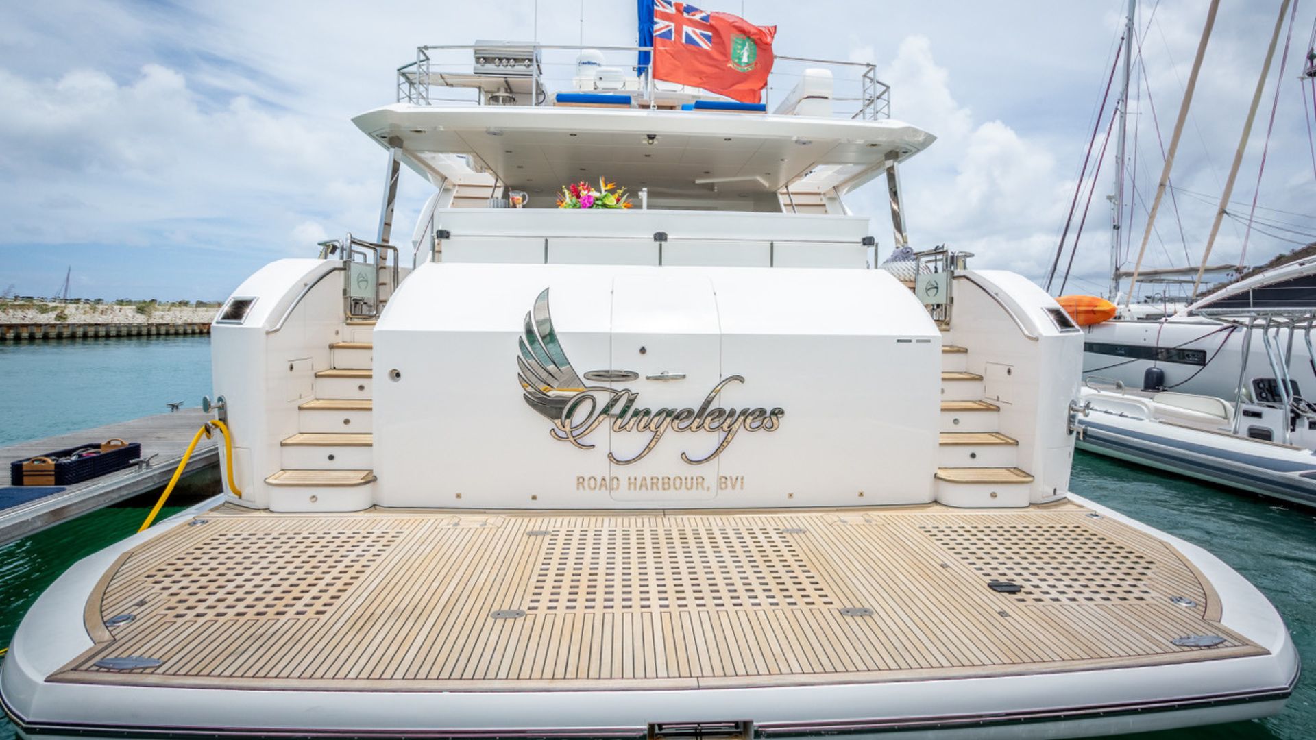 A luxury yacht named Angel Eyes docked at a marina, with a wooden deck and a British Virgin Islands flag flying at the back. The water and other boats are visible in the background.