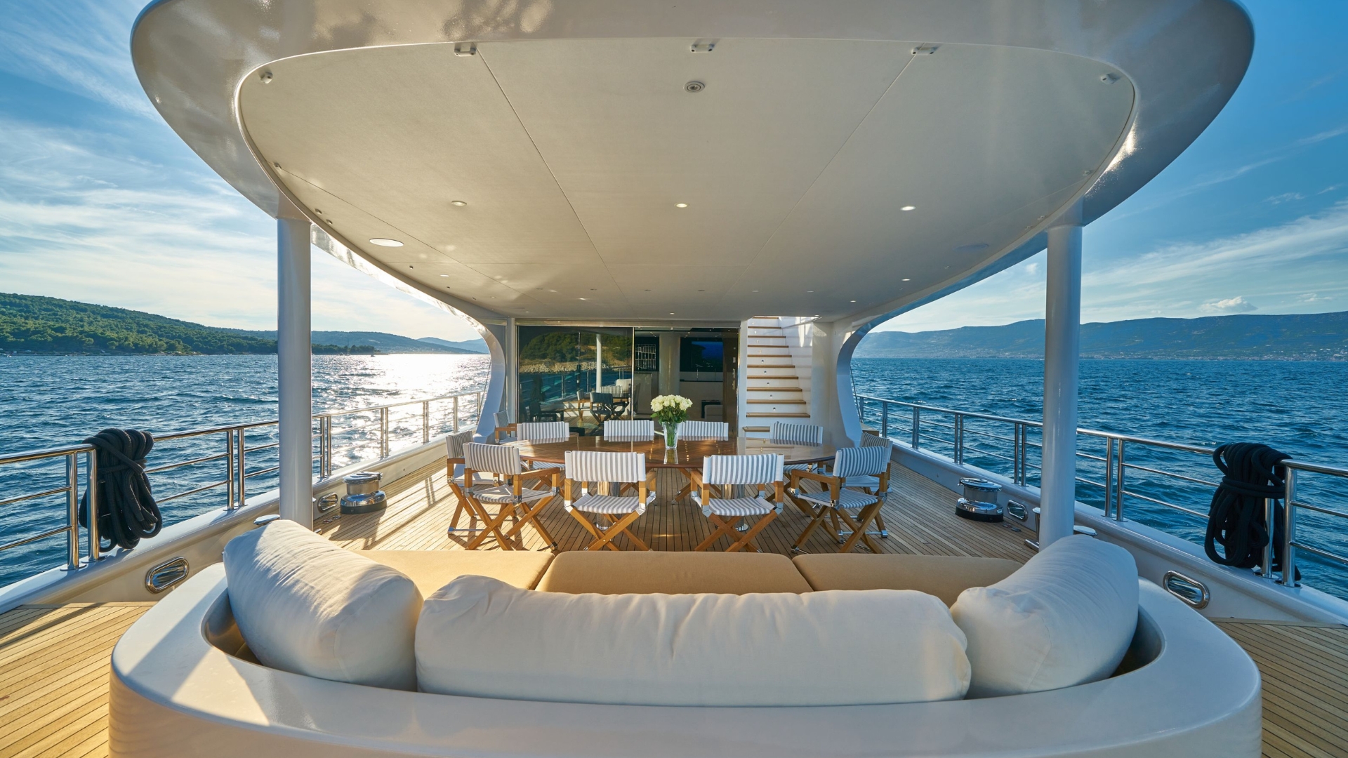 A luxury yacht deck with a white sofa, a table surrounded by chairs, and a view of calm blue sea and distant green hills under a sunny sky.