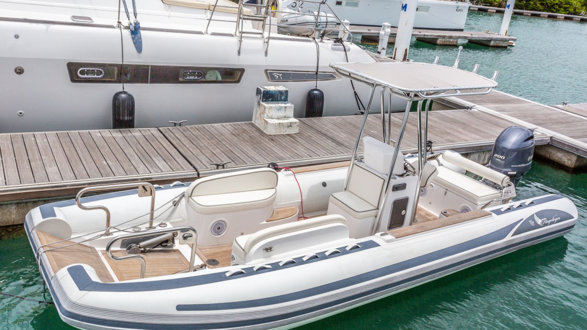 A white and gray inflatable motorboat with a T-top roof is docked beside a wooden pier, with a larger yacht and green water in the background.