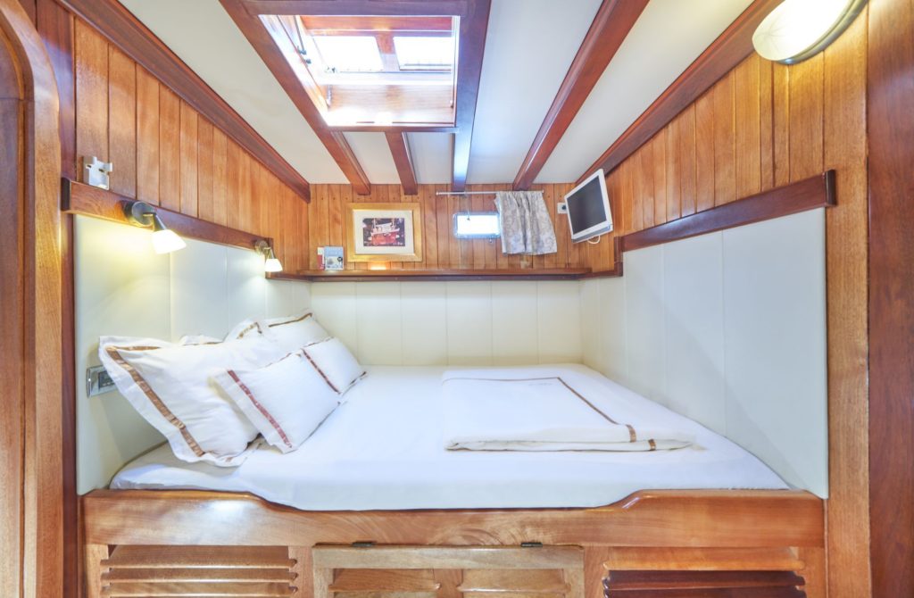LIBRA A cozy boat cabin with wood-paneled walls, a double bed with white linens, reading lights, a small window, wall-mounted TV, and a skylight above, creating a bright and inviting atmosphere.