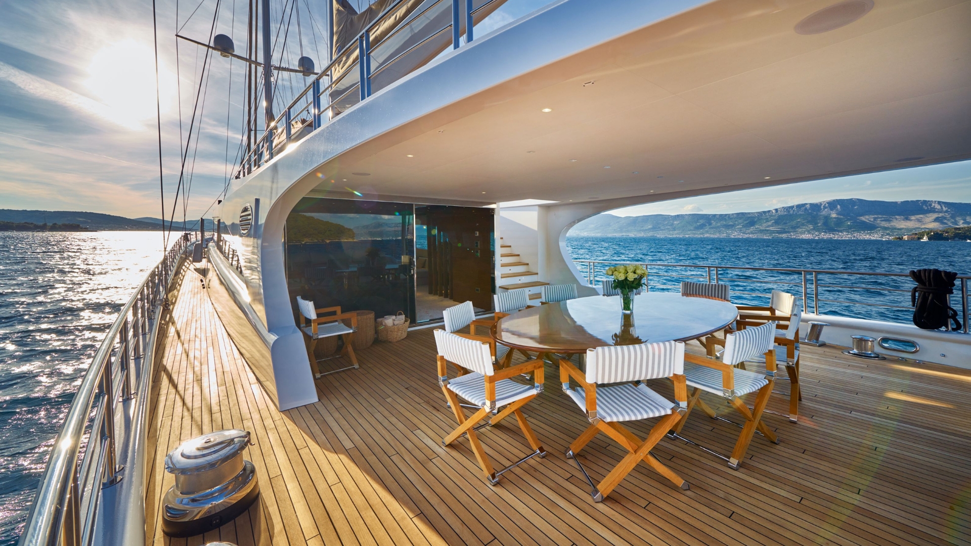 A luxurious yacht deck with a round dining table, striped chairs, and a vase of flowers, overlooking the sea with mountains in the distance under a bright, sunny sky.