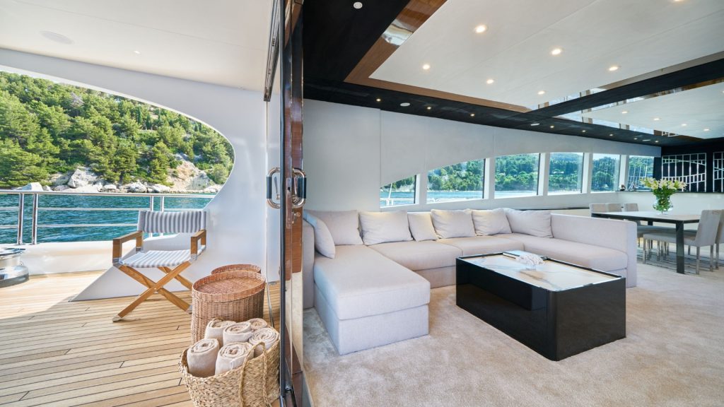 A luxury yacht interior with a spacious white sectional sofa, a black coffee table, and large windows overlooking water and green hills. Outside, a chair and basket with rolled towels sit on a wooden deck.