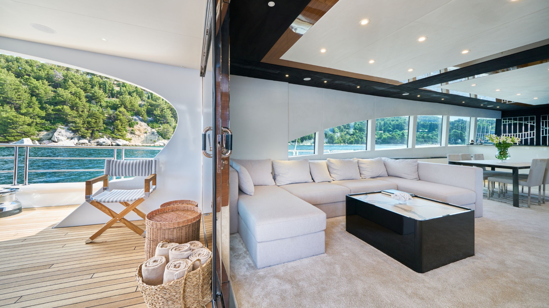 A luxury yacht interior with a spacious white sectional sofa, a black coffee table, and large windows overlooking water and green hills. Outside, a chair and basket with rolled towels sit on a wooden deck.