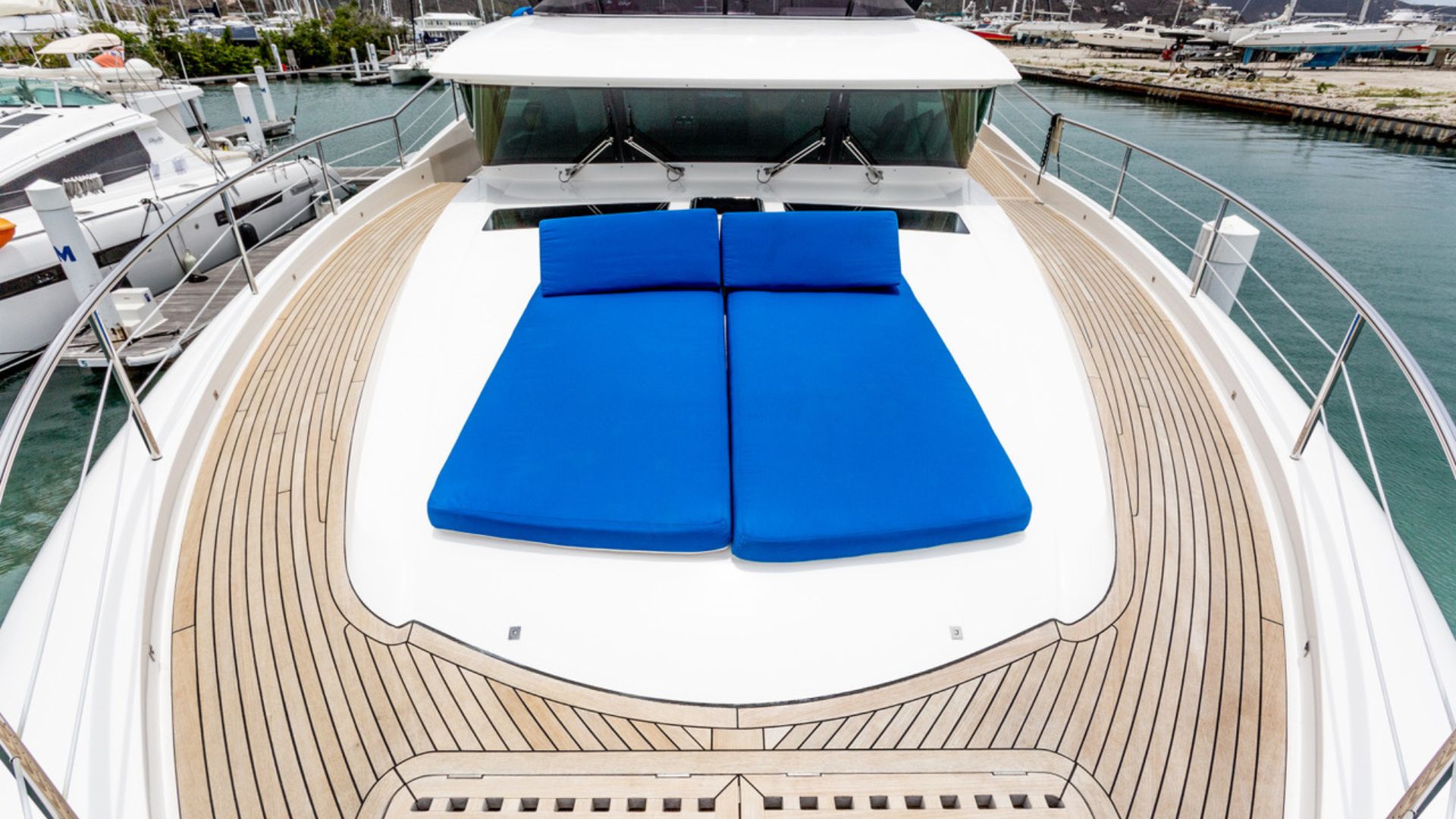The image shows the deck of a yacht with two bright blue lounge cushions placed side by side at the front, surrounded by light wooden flooring and railings, with a marina and other boats visible in the background.