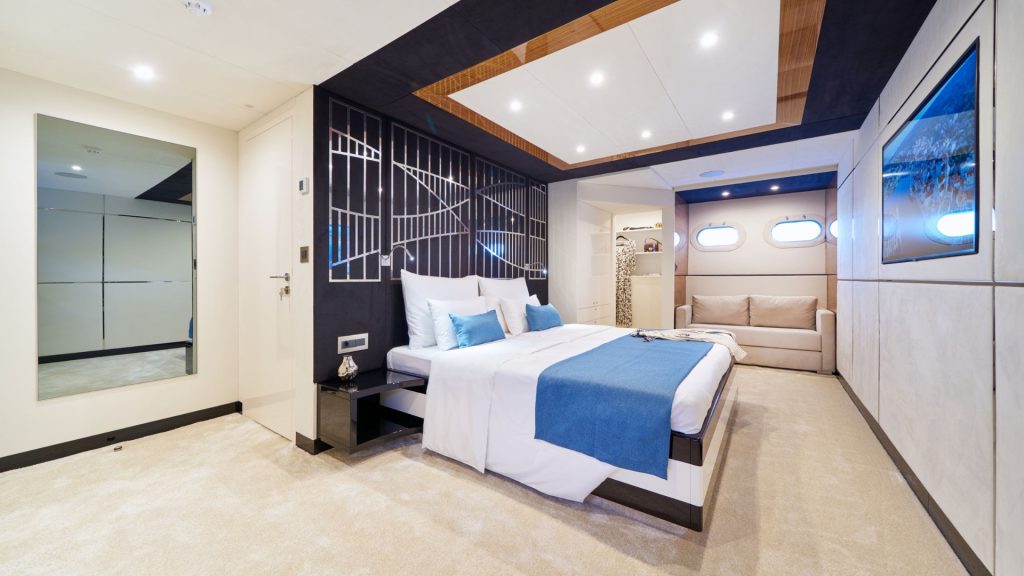 Modern yacht bedroom with a large bed, blue and white bedding, a beige sofa, mirrored closet, and porthole windows. The room has a beige carpet and sleek, contemporary decor with recessed lighting.