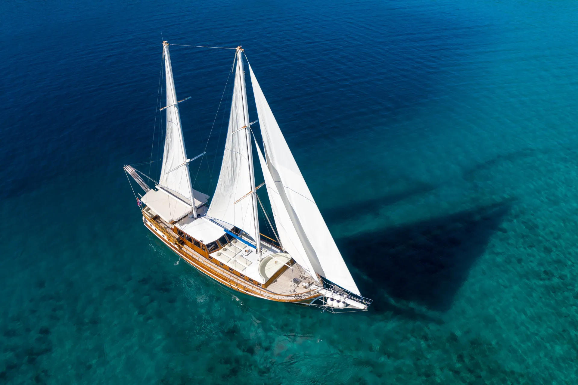 ANDI STAR Aerial view of a wooden sailboat with white sails gliding over clear blue water, casting a shadow on the sea below. The boat is surrounded by calm, turquoise ocean.