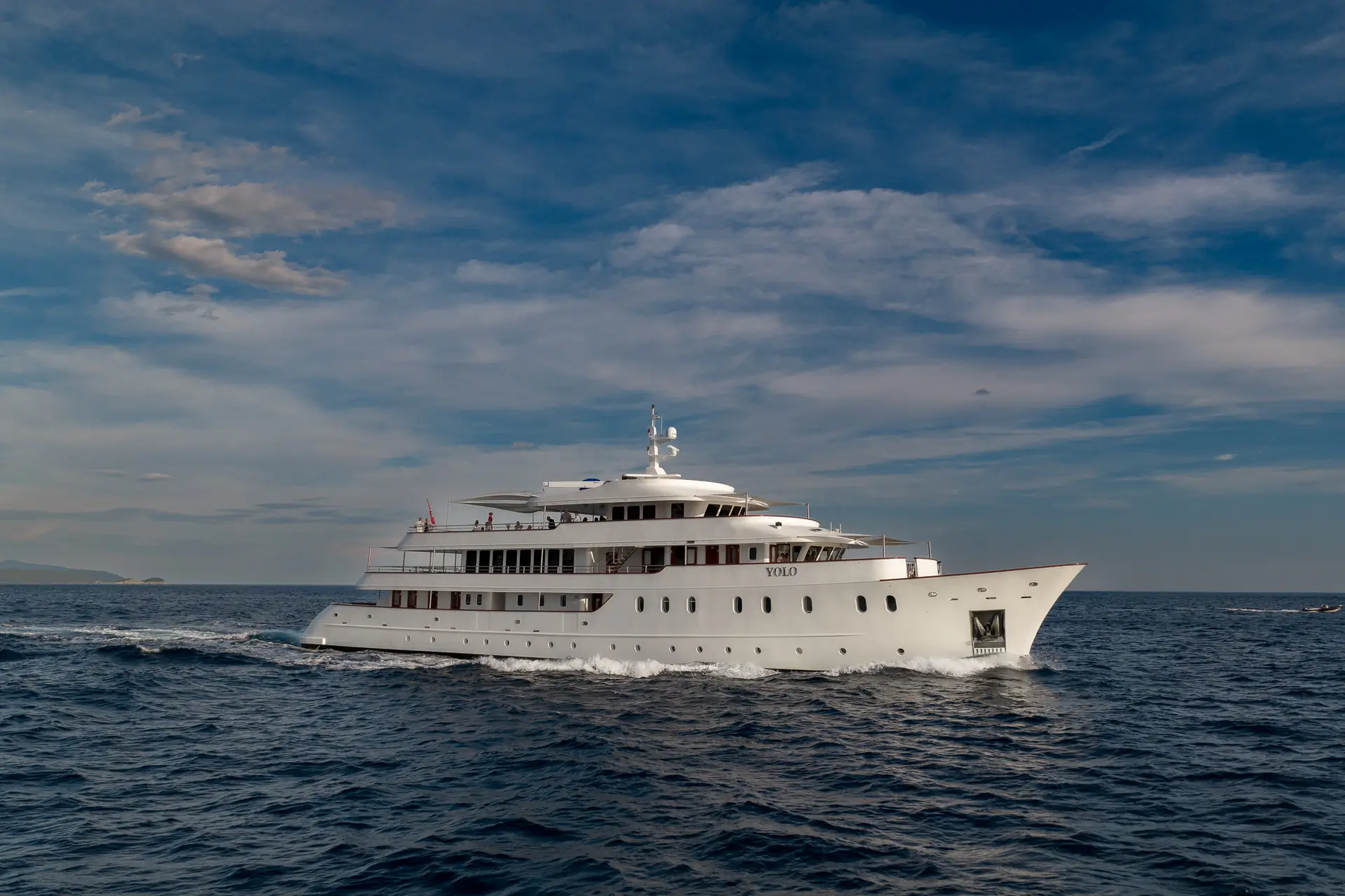 A large white luxury yacht cruises across deep blue ocean water under a partly cloudy sky, creating a small wake as it moves forward.