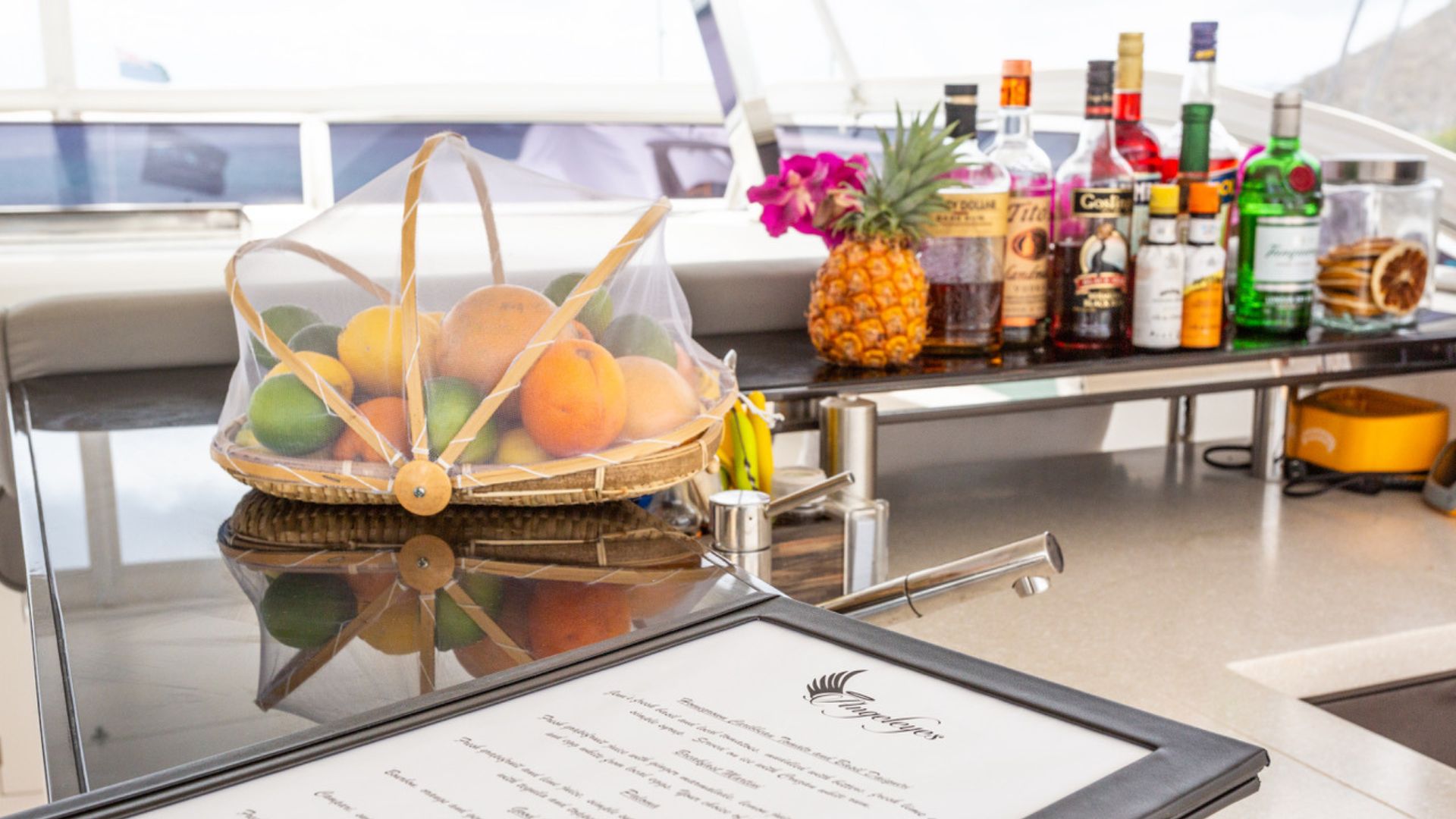 A basket of assorted fruit under a mesh cover sits on a counter next to bottles of liquor, a pineapple, and a pink flower arrangement, with a framed menu in the foreground on a yacht.