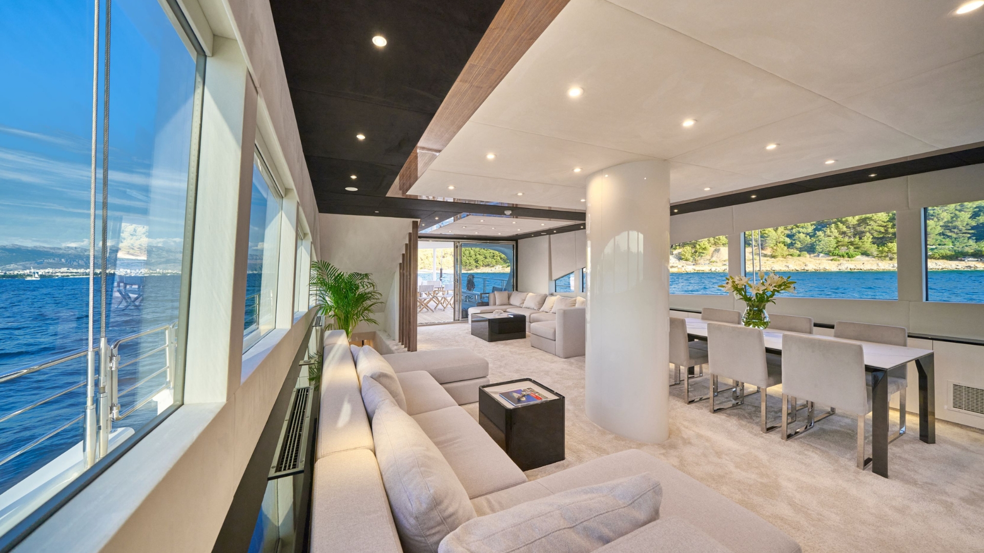 A spacious, modern yacht interior with large windows showing ocean views, a cozy lounge area with light gray sofas, a dining table with white chairs, and natural light streaming in.