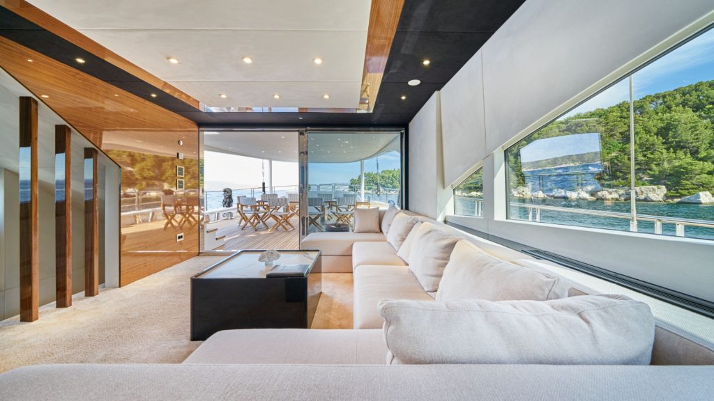 Modern yacht interior with large beige sectional sofa, sleek wooden accents, and floor-to-ceiling windows offering scenic views of water and greenery. Outdoor dining area visible through glass doors.
