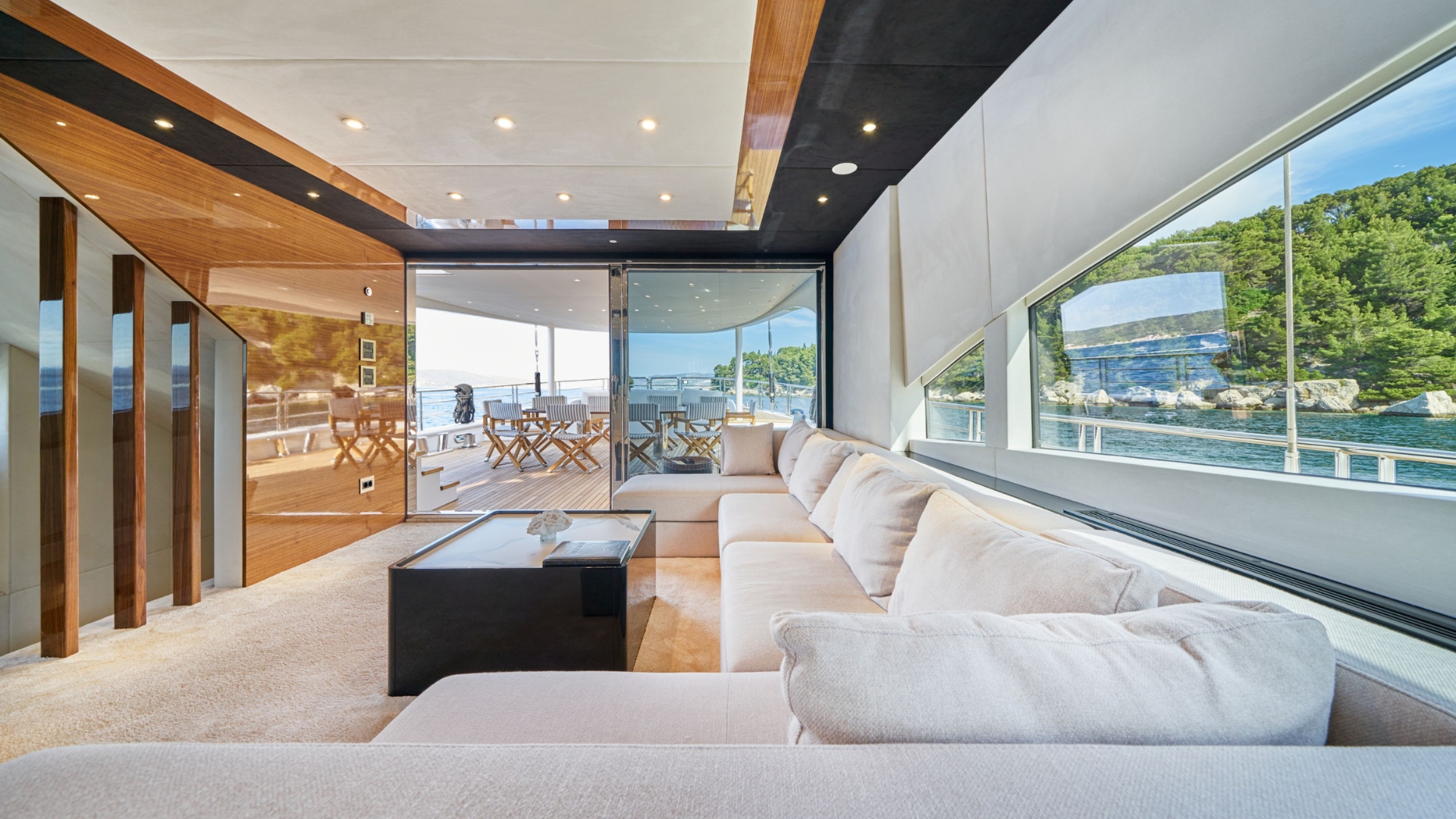 Modern yacht interior with large beige sectional sofa, sleek wooden accents, and floor-to-ceiling windows offering scenic views of water and greenery. Outdoor dining area visible through glass doors.