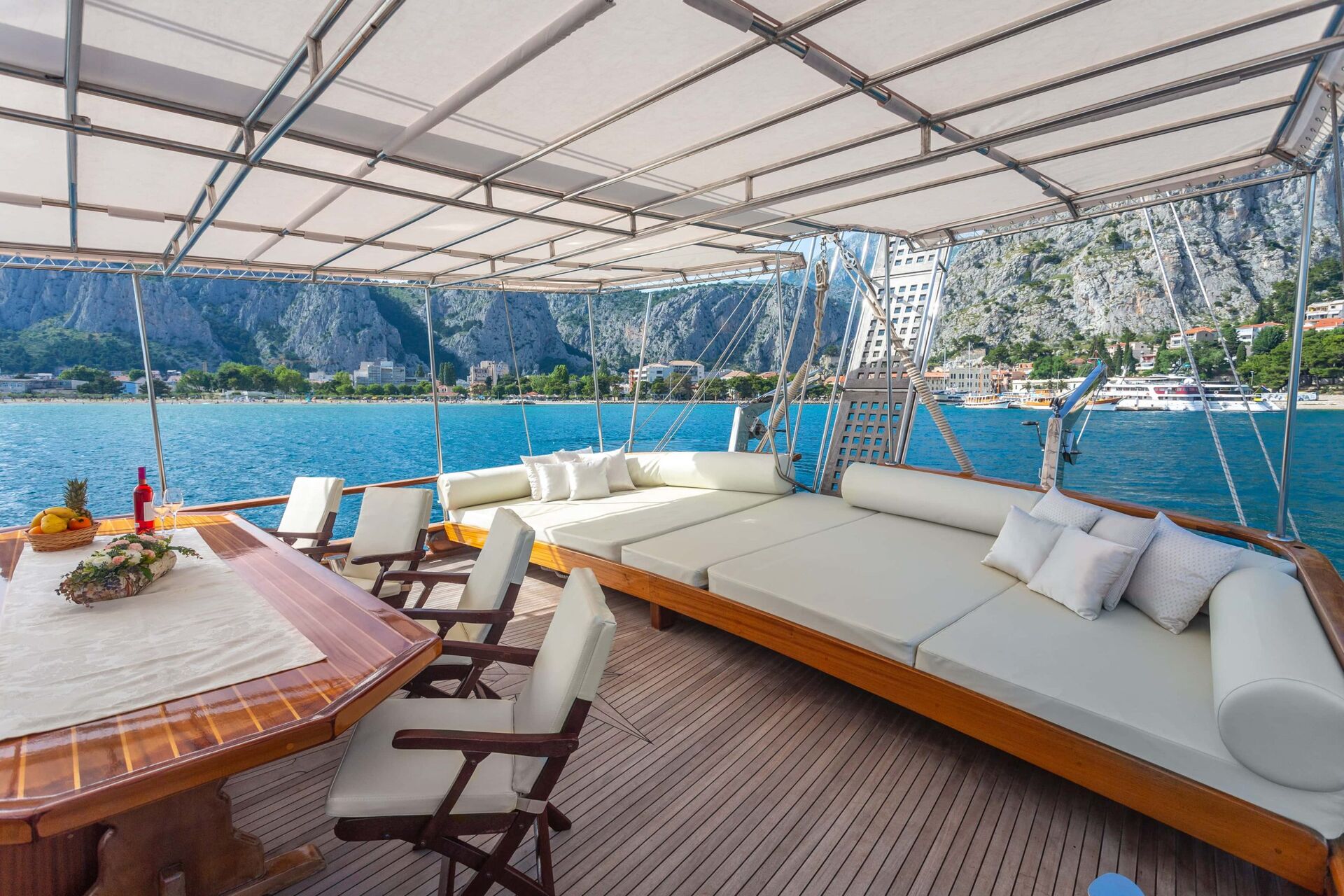 A yacht’s deck with cushioned seating, a wooden dining table set for a meal, and chairs, overlooks calm blue water and a coastal town with mountains in the background under a canopy.