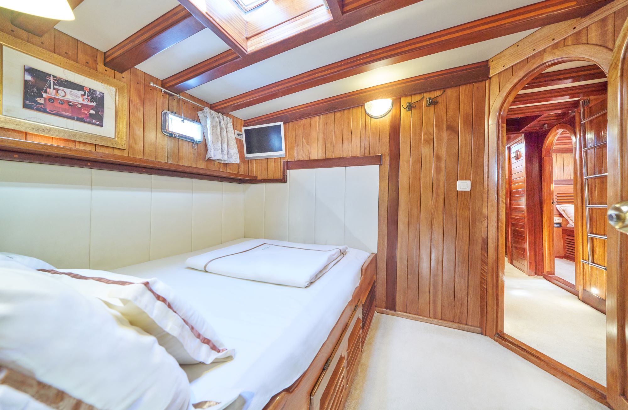 LIBRA A cozy cabin with wooden walls and ceiling, a neatly made bed, a small TV, a framed picture, and an arched doorway leading to another room, illuminated by natural and artificial light.