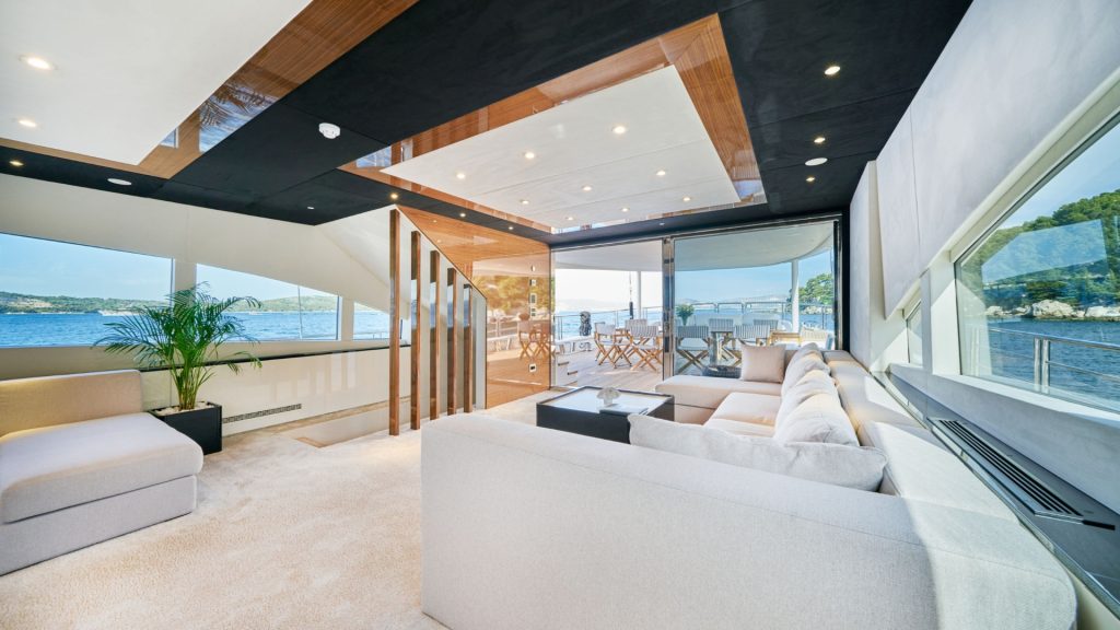 Modern yacht interior with large cream sectional sofa, sleek wood and black ceiling accents, bright lighting, a potted plant, and floor-to-ceiling windows showcasing views of water and outdoor deck seating.