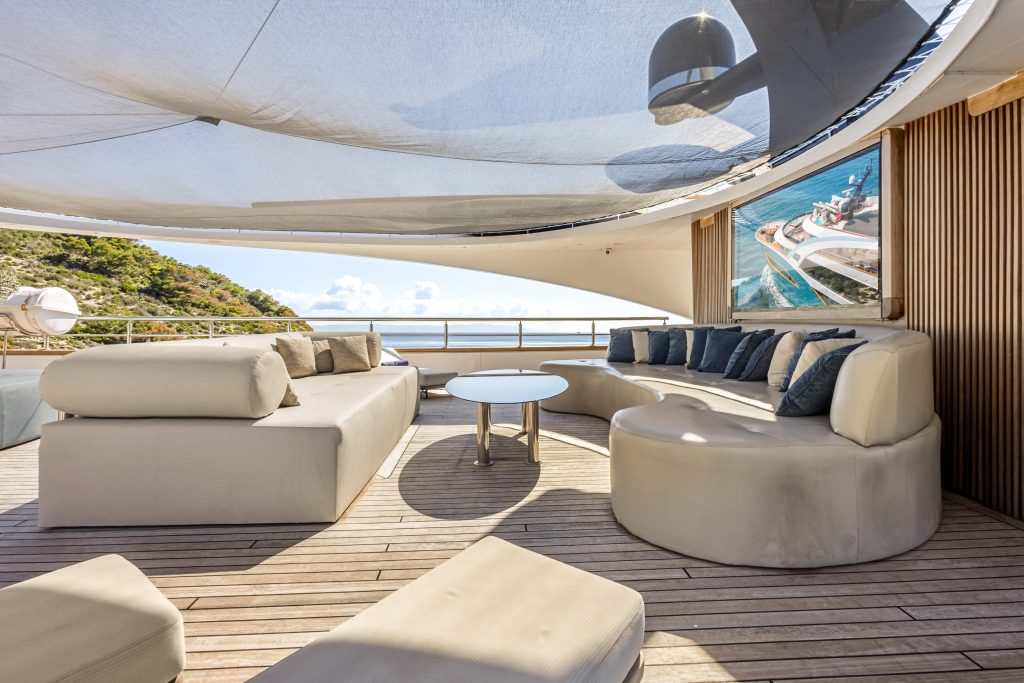 Luxury yacht deck with modern curved sofas, cushions, and a round table on wooden flooring. A large canopy provides shade. Ocean and cliffs are visible in the background on a sunny day.