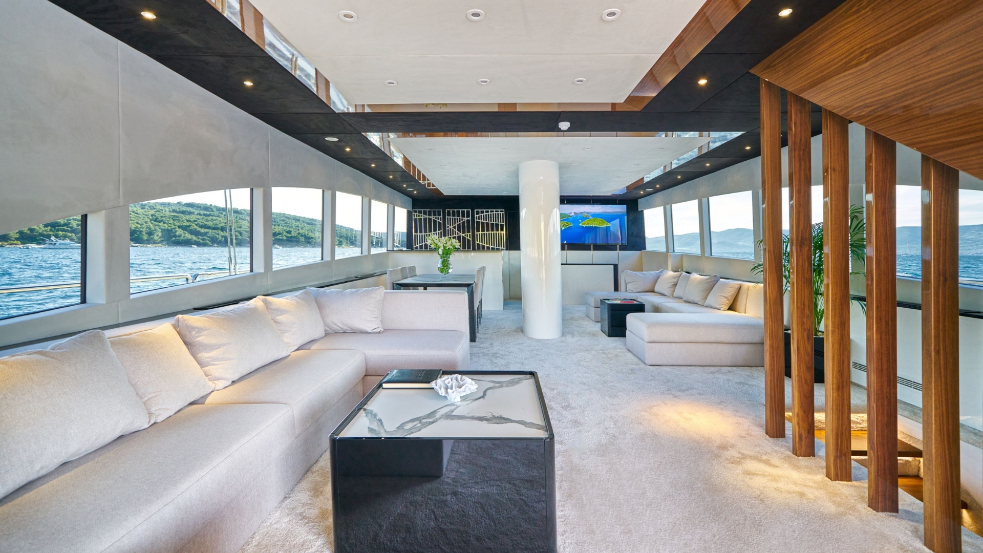 Luxurious yacht interior with large beige sofas, modern tables, wood accents, carpeted floor, and panoramic windows showing water and green hills outside; a TV is mounted on the far wall.