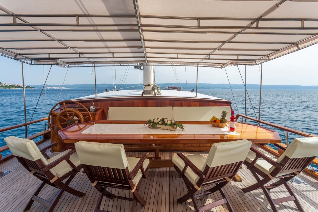 A wooden dining table with cushioned chairs is set on the deck of a yacht, overlooking calm blue sea under a canopy. A flower arrangement and fruit plate decorate the table. The coastline is visible in the distance.