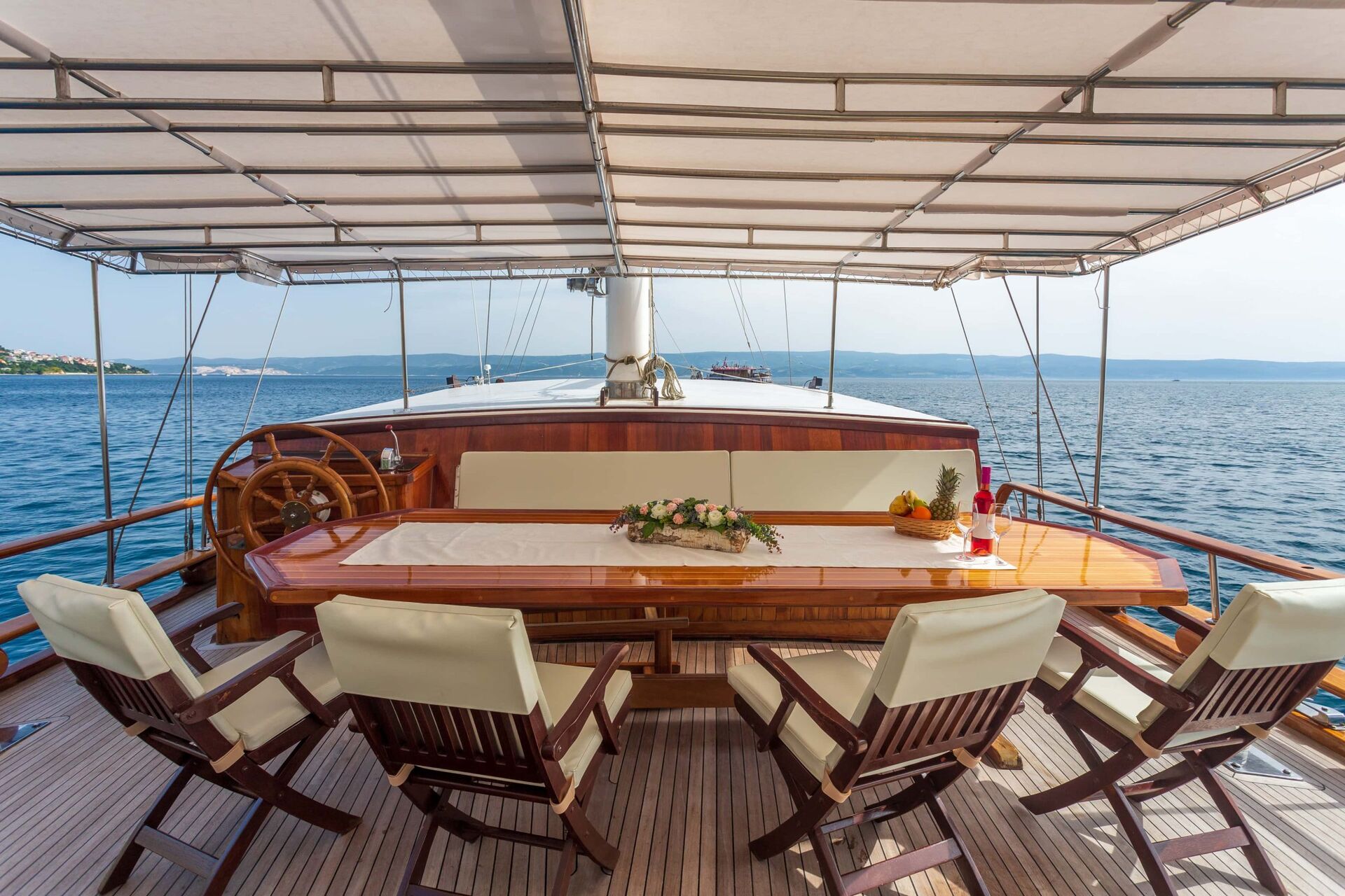 A wooden dining table with cushioned chairs is set on the deck of a yacht, overlooking calm blue sea under a canopy. A flower arrangement and fruit plate decorate the table. The coastline is visible in the distance.