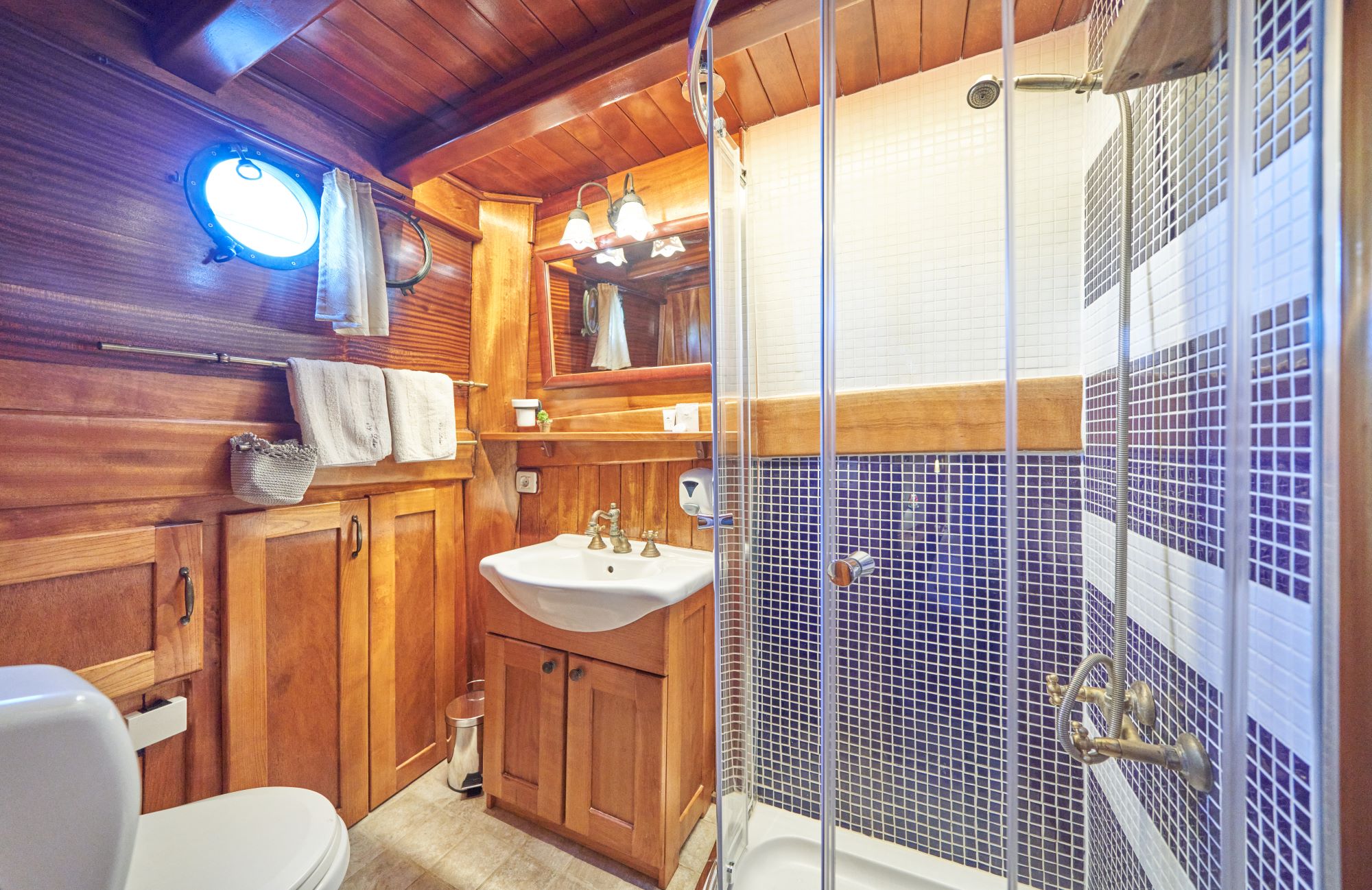 LIBRA A cozy bathroom with wood-paneled walls and ceiling, a small sink with a wooden cabinet, a glass shower with mosaic tiles, a round window, white towels hanging, and warm lighting.