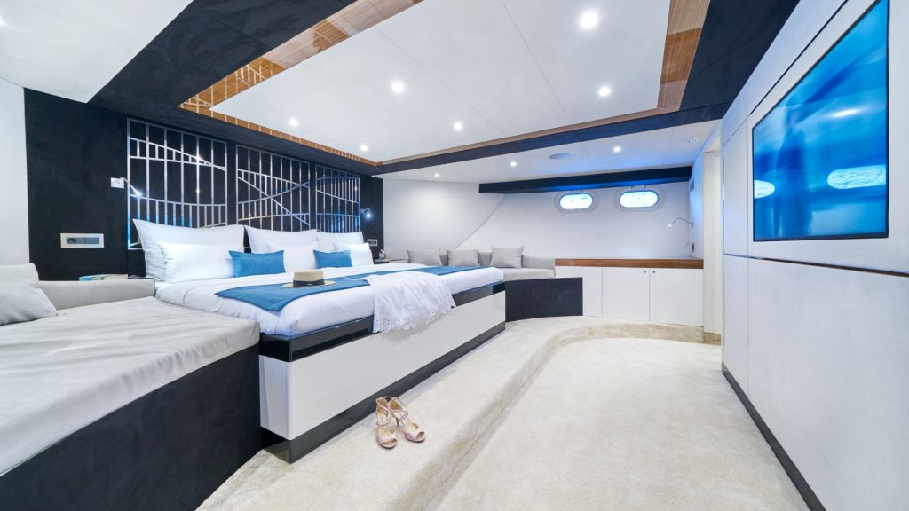 A modern, luxurious yacht bedroom with a large bed, soft white bedding, built-in sofa seating, a wall-mounted TV, and two small porthole windows. A pair of high heel shoes are on the cream carpet floor.