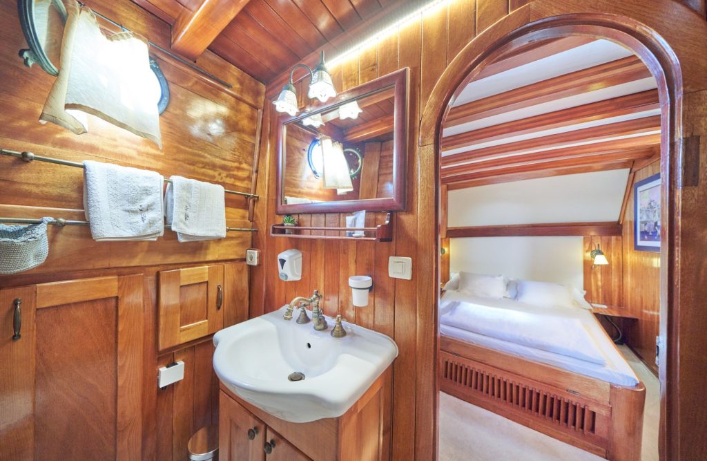 LIBRA A cozy, wood-paneled bathroom with a white sink and towels, next to an arched doorway leading to a bedroom with a white bed and wooden beams on the ceiling. Natural light enters through porthole windows.