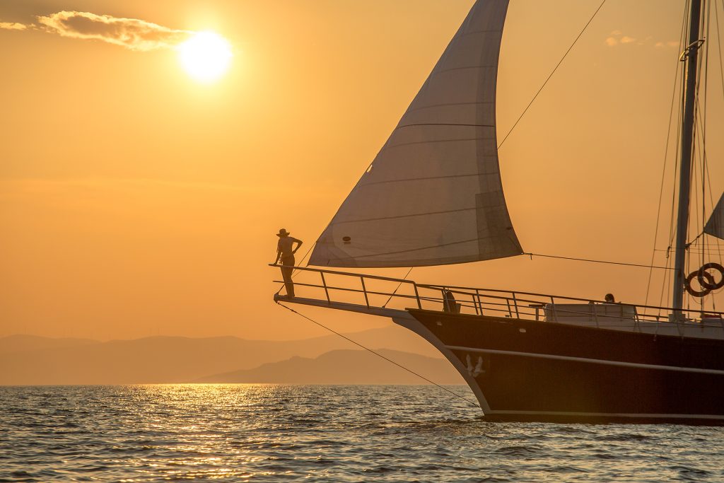A person stands on the bow of a sailboat at sunset, with the sun low in the sky, casting a warm orange glow over the calm sea and distant hills—an ideal moment to enjoy a yacht charter as the water reflects the sunlight.