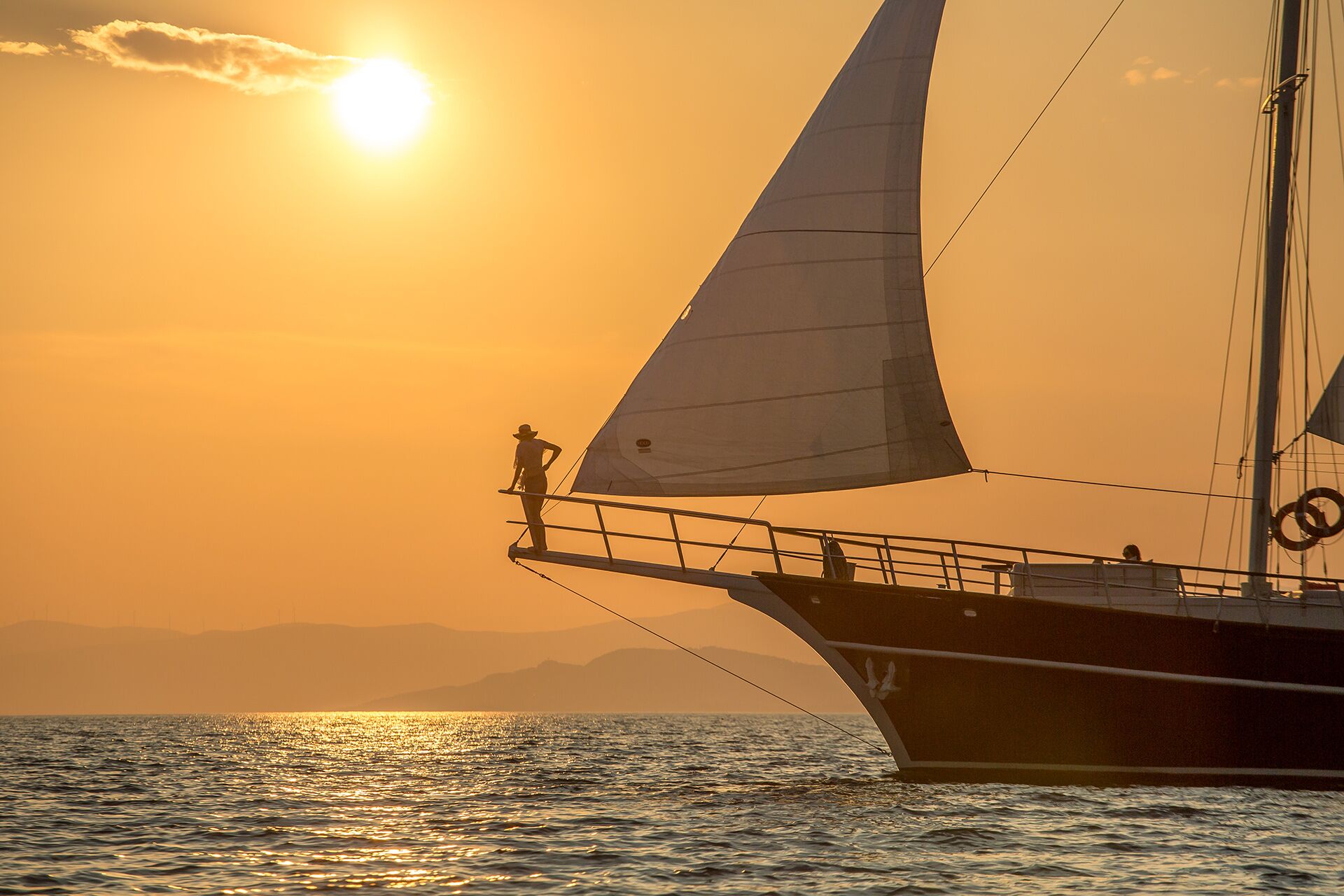 A person stands on the bow of a sailboat at sunset, with the sun low in the sky, casting a warm orange glow over the calm sea and distant hills—an ideal moment to enjoy a yacht charter as the water reflects the sunlight.
