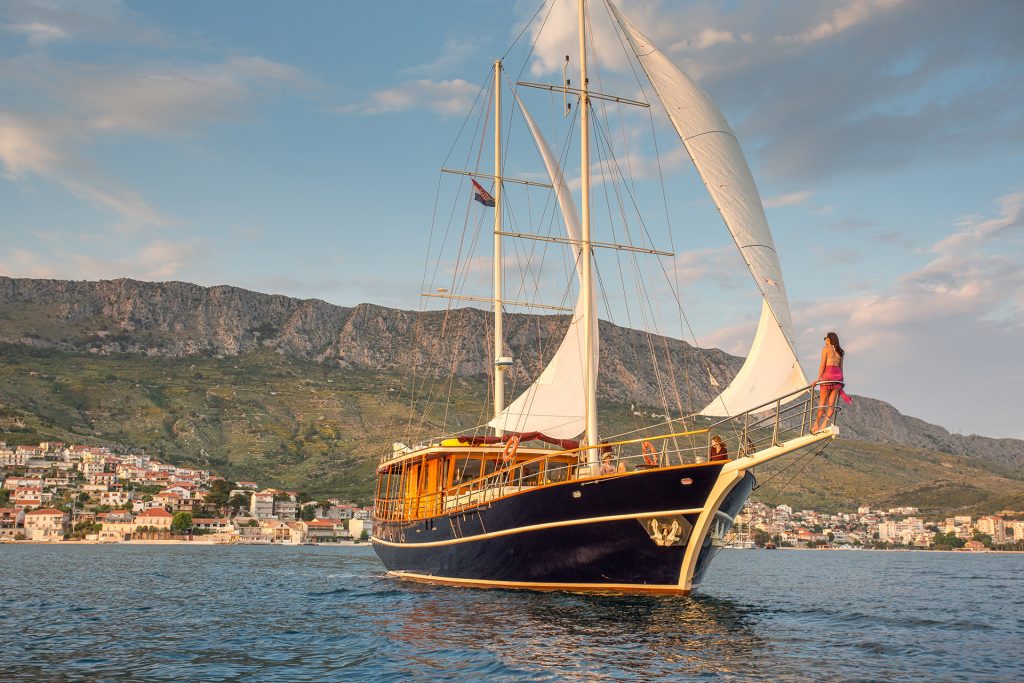A large sailboat with white sails glides on calm water near a coastal town, with mountains in the background. Perfect for yacht charter, a woman in a red dress stands at the bow, looking toward the horizon.
