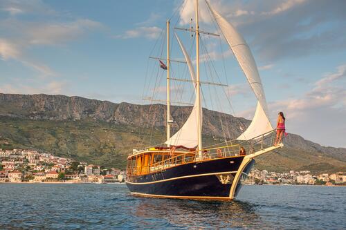 A large sailboat with white sails glides on calm water near a coastal town, with mountains in the background. Perfect for yacht charter, a woman in a red dress stands at the bow, looking toward the horizon.