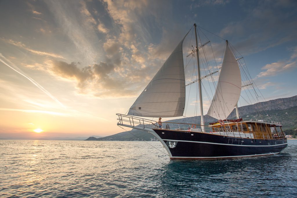 A large sailboat with white sails glides on calm water at sunset, with mountains and a partly cloudy sky in the background—perfect for an unforgettable yacht charter experience.