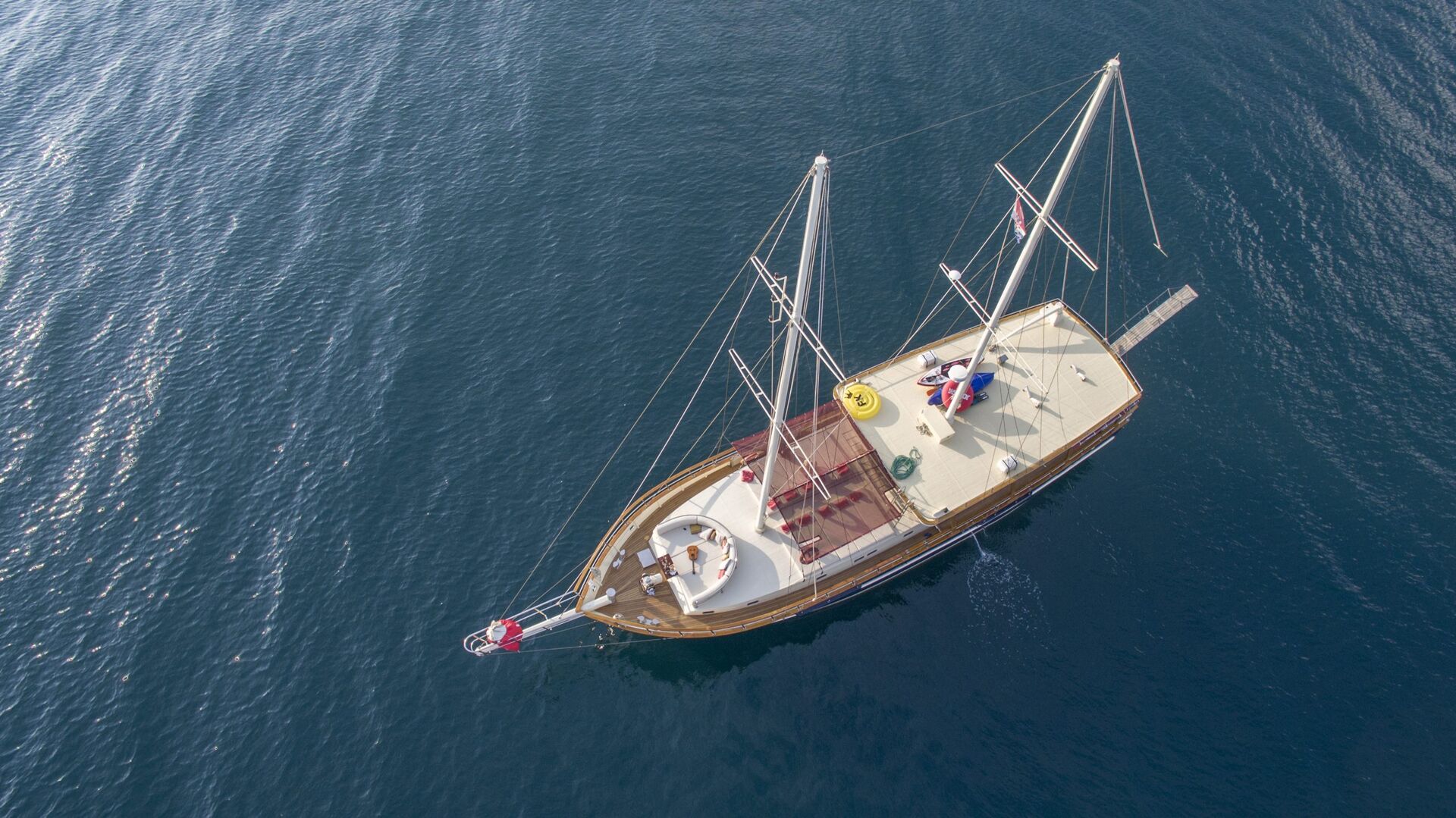 Aerial view of a sailboat with a wooden deck and two masts, floating on calm, deep blue water—a perfect scene for those seeking yacht rental or yachts for charter. A few people relax on deck as the boat’s wake trails behind it.