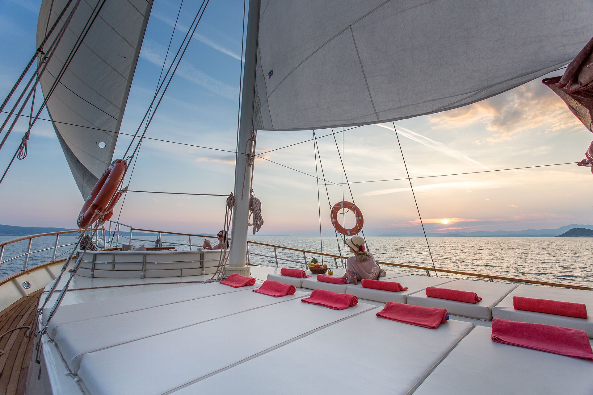 A person relaxes on the deck of a sailing yacht at sunset, surrounded by neatly rolled pink towels, with calm sea and distant mountains under a partly cloudy sky.