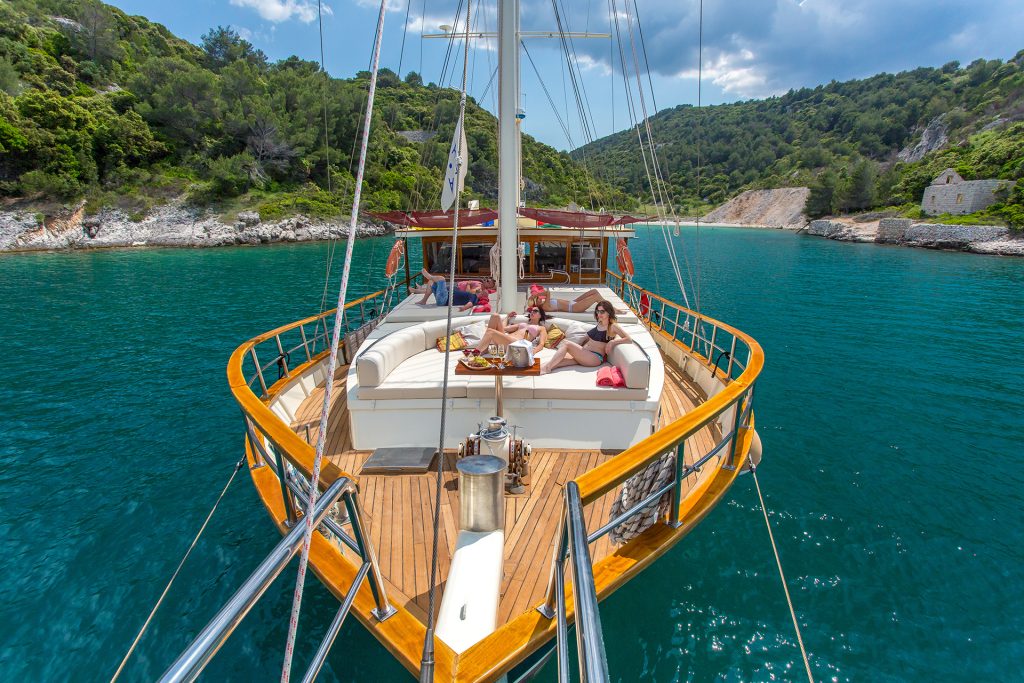 A group of people relax on the deck of a luxury yacht charter anchored in a calm, turquoise bay surrounded by green hills and rocky shores under a partly cloudy sky.