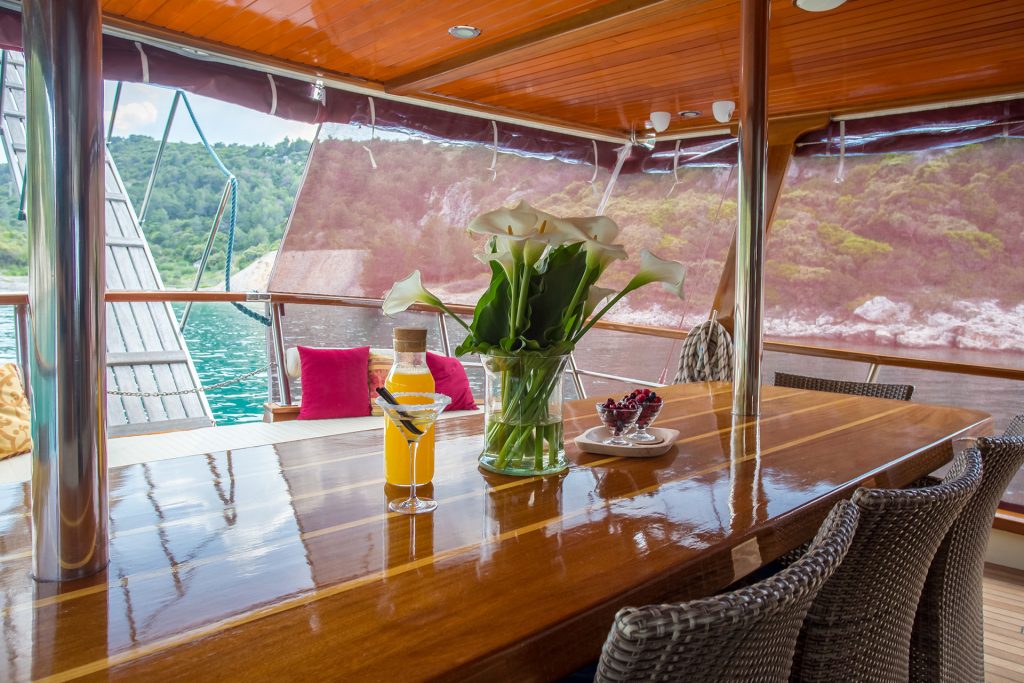A polished wooden table on a yacht charter deck, set with a vase of white flowers, two drinks, and a plate of berries. Cushioned seats invite you to relax and enjoy clear views of green hills and sparkling water.