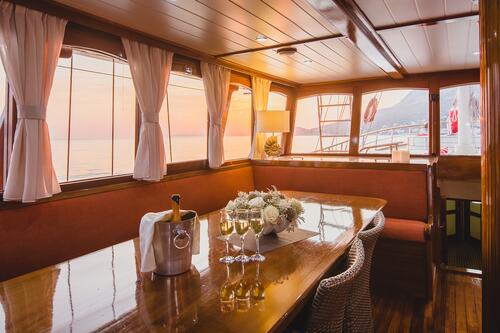 A cozy yacht charter dining area with a polished wooden table, four glasses of white wine, a flower arrangement, and a champagne bottle in an ice bucket. Large windows reveal a sunset over calm water outside.
