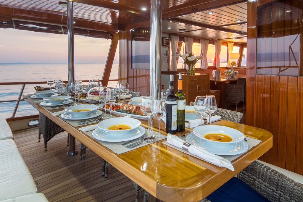 A set dining table with soup, wine, and glasses on a yacht’s wooden deck overlooks the calm sea at sunset—perfect for those seeking a luxurious yacht rental, with elegant tableware and cozy indoor seating visible in the background.