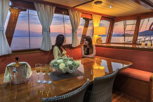 Two women sit and talk at a polished wooden table with flowers and champagne inside a cozy yacht charter cabin at sunset, with large windows revealing the sea and distant mountains.