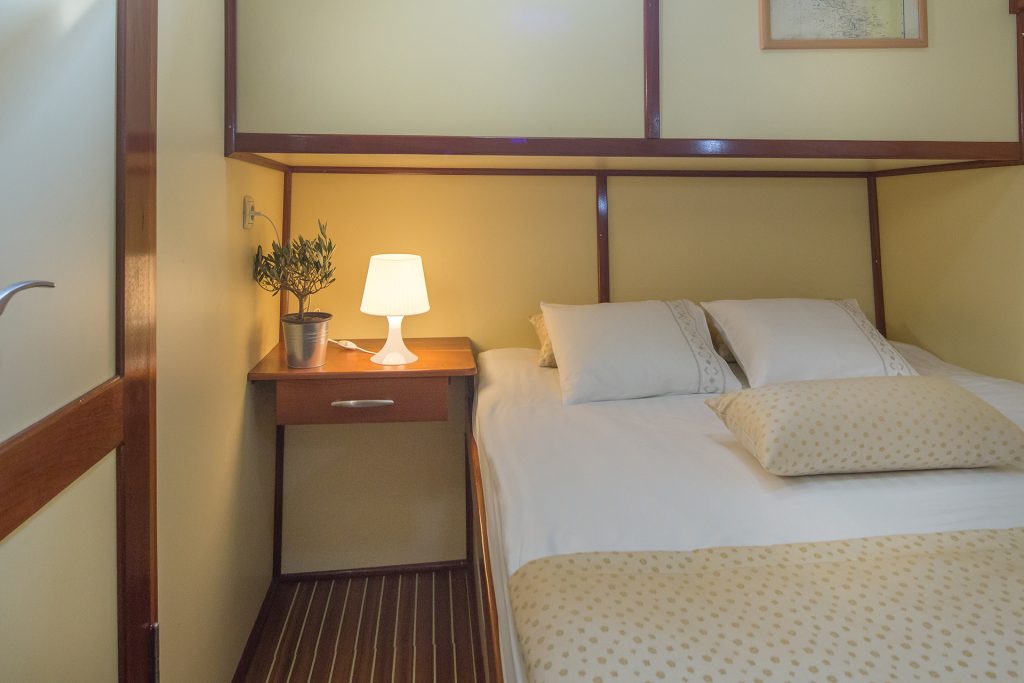 A cozy bedroom on a luxury yacht charter, featuring a neatly made double bed with a polka-dot blanket, two pillows, a nightstand with a potted plant, and a lit lamp—all set against light yellow walls and elegant wood accents.