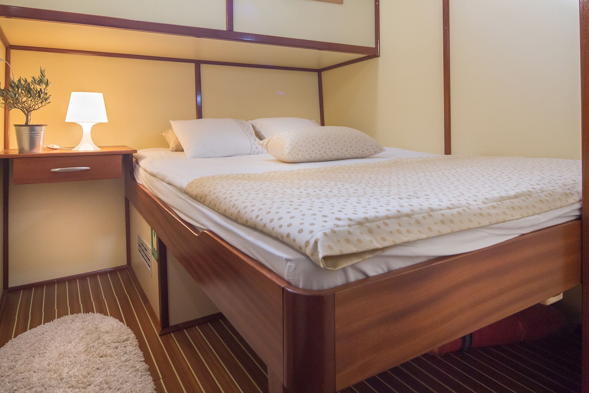 A cozy bedroom with a neatly made bed in cream and beige, a small nightstand with a lamp and potted plant, and a soft white rug on wooden floors. The warm lighting and wooden furniture give it the inviting feel of a private yacht charter retreat.